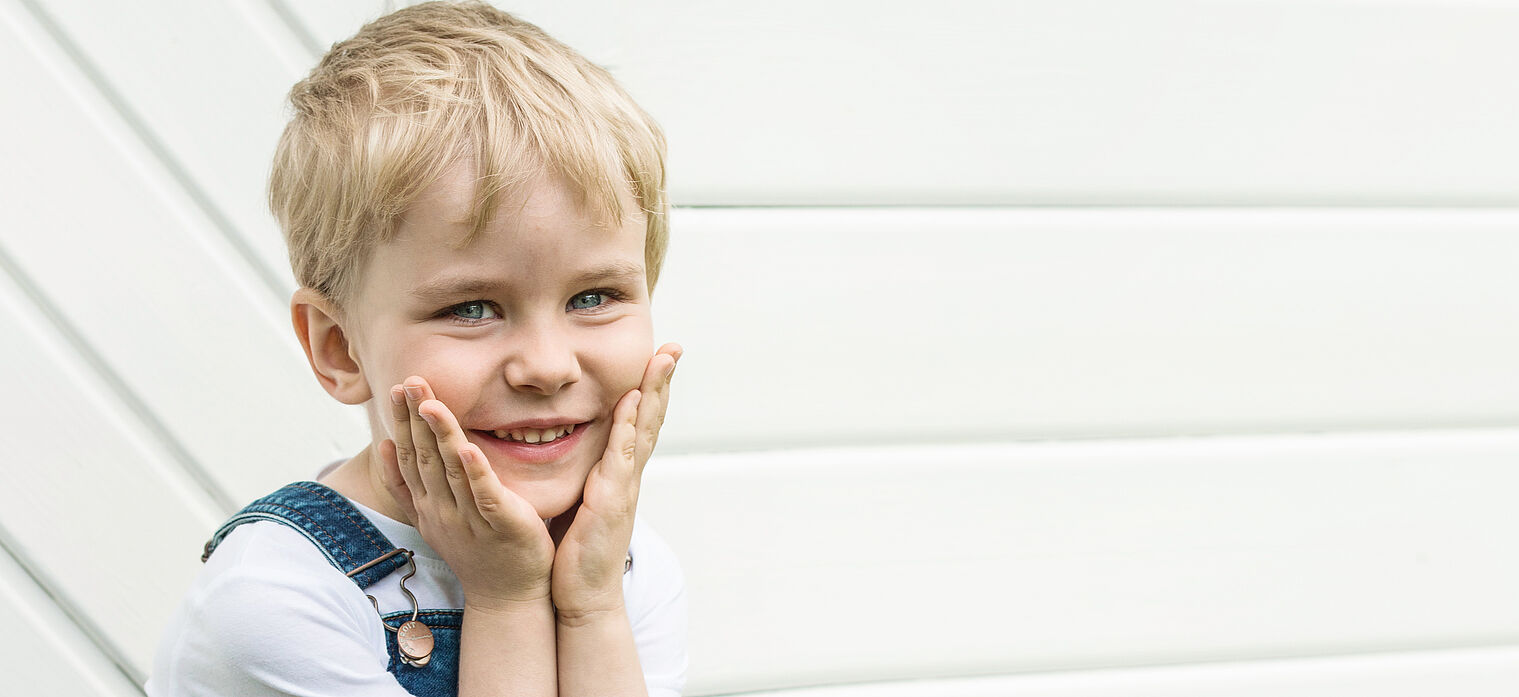 A young boy with blond hair smiling, holding his cheeks with both hands, standing in front of a white background.